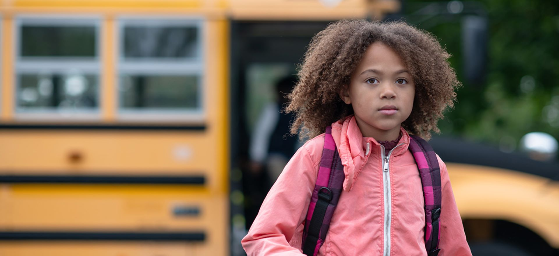 A school-age child, wearing a backpack and a pink jacket, stands in front of a school bus and stares at the camera with a serious expression. 